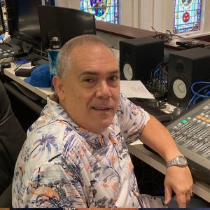 A staff member at the audio-visual console in the First United Methodist Church of Sarasota sanctuary, with stained glass visible in the background.